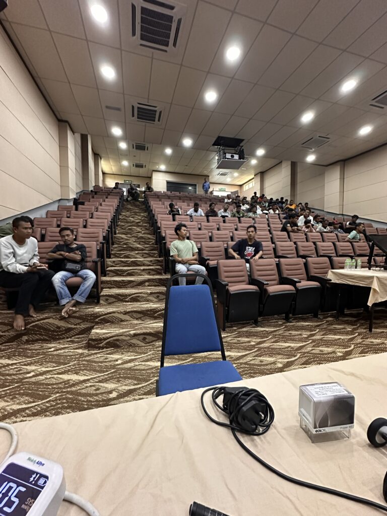 Pekerja duduk di kerusi dewan auditorium menunggu giliran pemeriksaan, dengan alat tekanan darah di atas meja hadapan