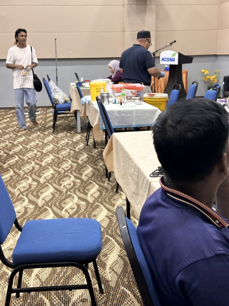 Stesen pemeriksaan kesihatan di dalam dewan dengan meja peralatan, kakitangan klinik dan pekerja